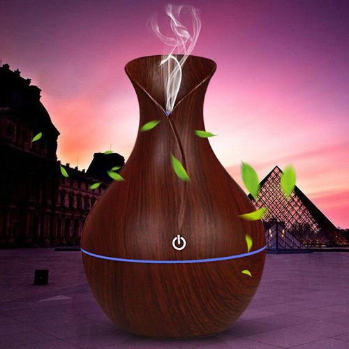 LED ultrasonic aroma humidifier and oil diffuser with wood grain vase design releasing mist and green leaves, set against an outdoor evening backdrop near modern architecture.