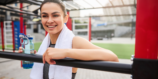 Smiling woman with towel and water bottle resting on outdoor gym bars showcasing compact wellness tech for fitness recovery and self-care